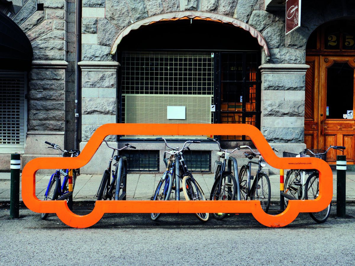 Ein Fahrradabstellplatz am Rand einer städtischen Straße, die an Häusern mit Arkadenbögen liegt. Vor den abgestellten 8 Rädern wurde straßenseitig ein oranger Rahmen gestellt, der etwa den Umriss eines Autos in Originalgröße hat. 