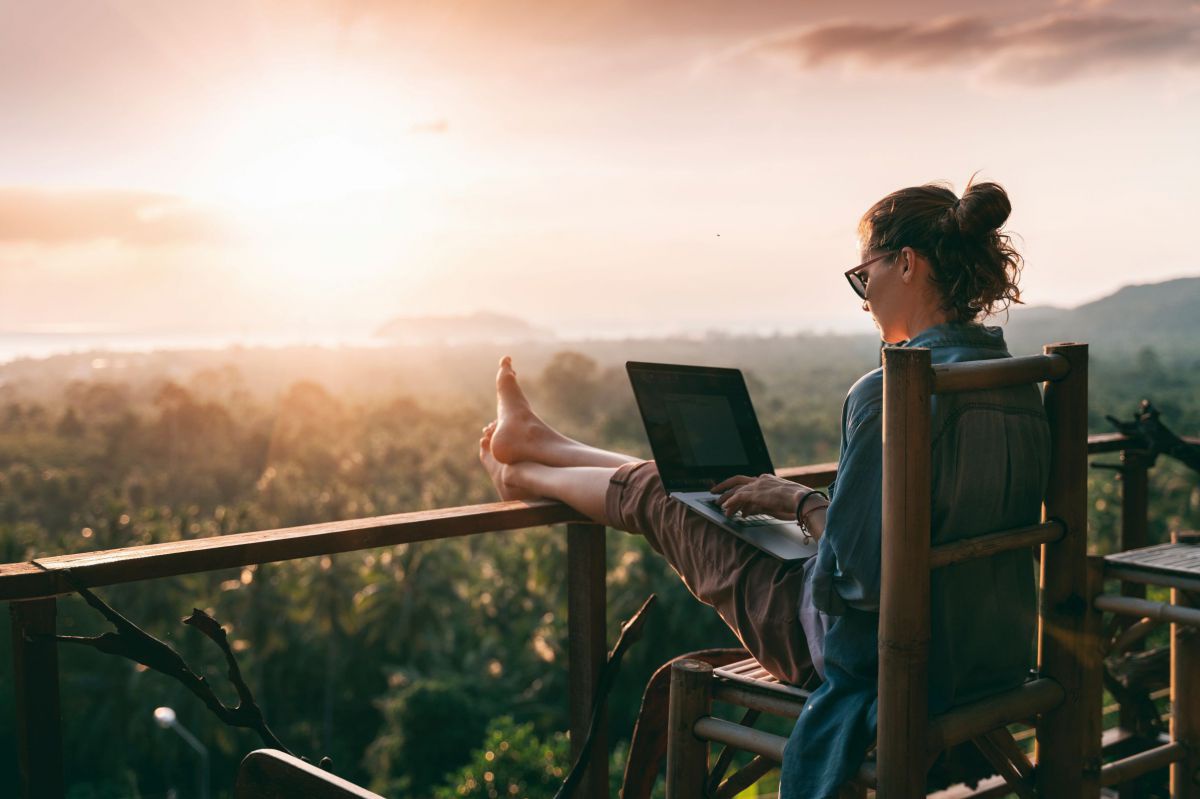 Auf einem Balkon sitzt eine Frau mit Sonnenbrille auf einem Bambusstuhl, die Beine am Geländer abgelegt, einen Laptop auf dem Schoß. Vor ihr erstreckt sich ein tropischer Wald, vom Licht der Abendsonne überflutet.