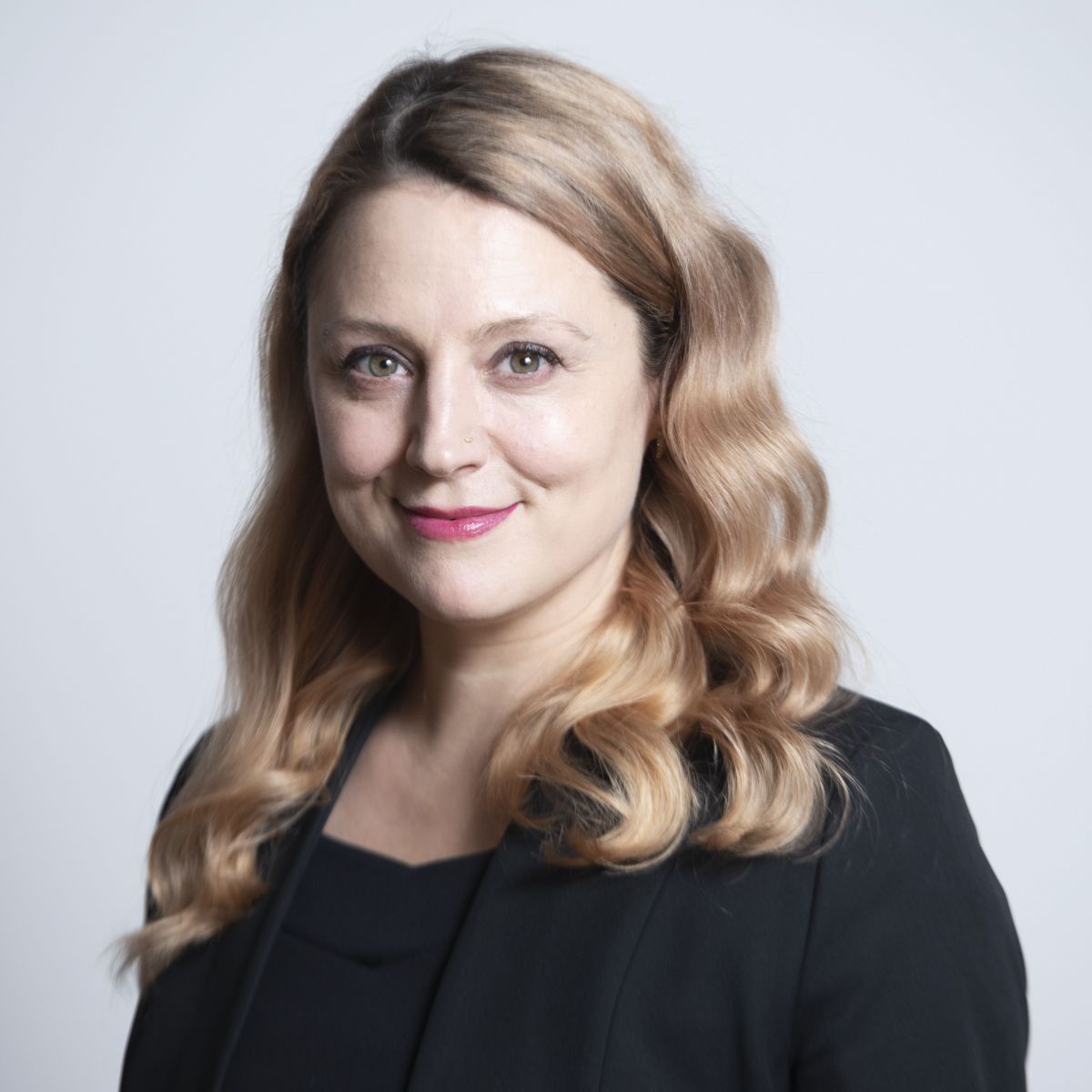 Clara Maria Schenk: Eine Frau mit heller Haut, blondem, gewelltem Haar im schwarzen Business-Anzug. Lächelt mit gechlossenen, roten Lippen, Blick direkt in die Kamera.