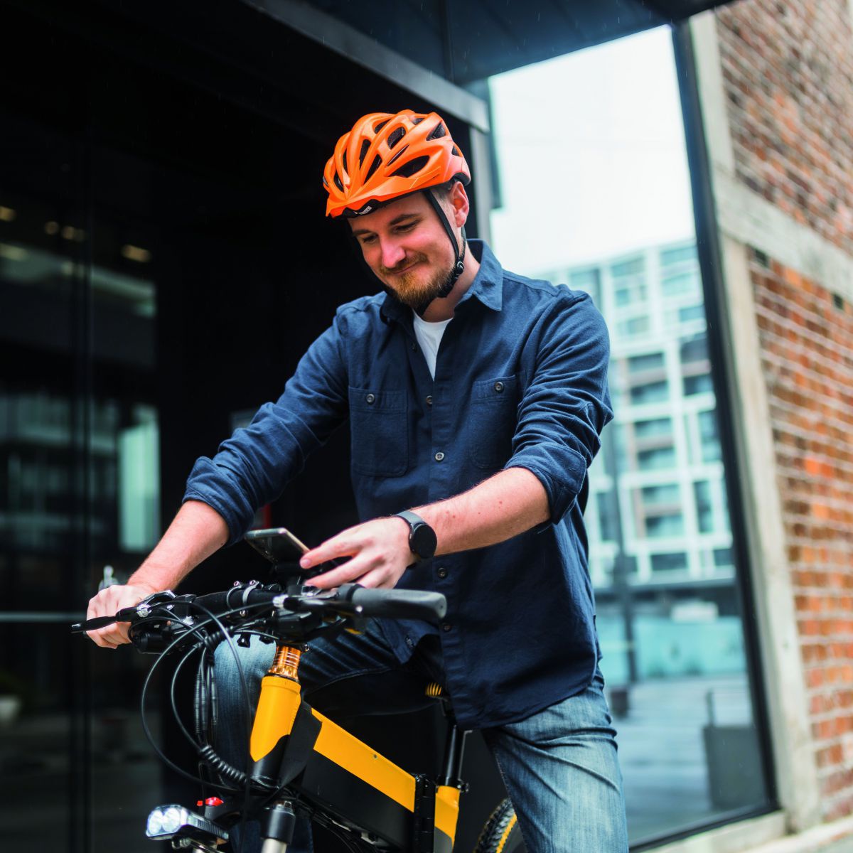 Ein Mann mit orangerotem Fahrradhelm, er sitzt auf dem Rad und hat gerade angehalten, ein Fuß am Boden abgestellt, liest etwas auf seinem Handy. Hinter ihm Backsteingebäude mit Glasfront.