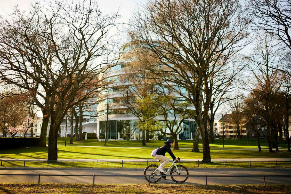 Radfahrer mit Helm, Pullover und Stoffhose, also keine Freizeit-Radfahrkleidung, der durch einen Park fährt. Das Bild steht symbolisch für Radpendler*innen