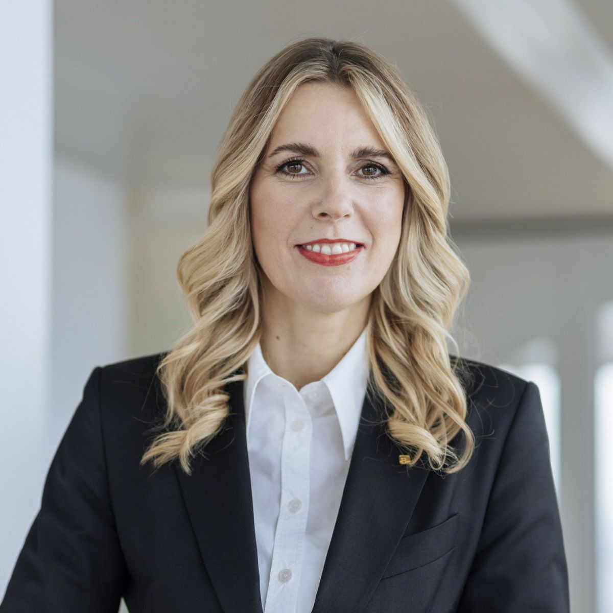 Frau mit blonden, welligen Haaren im schwarzen Business-Anzug und weißer Bluse.