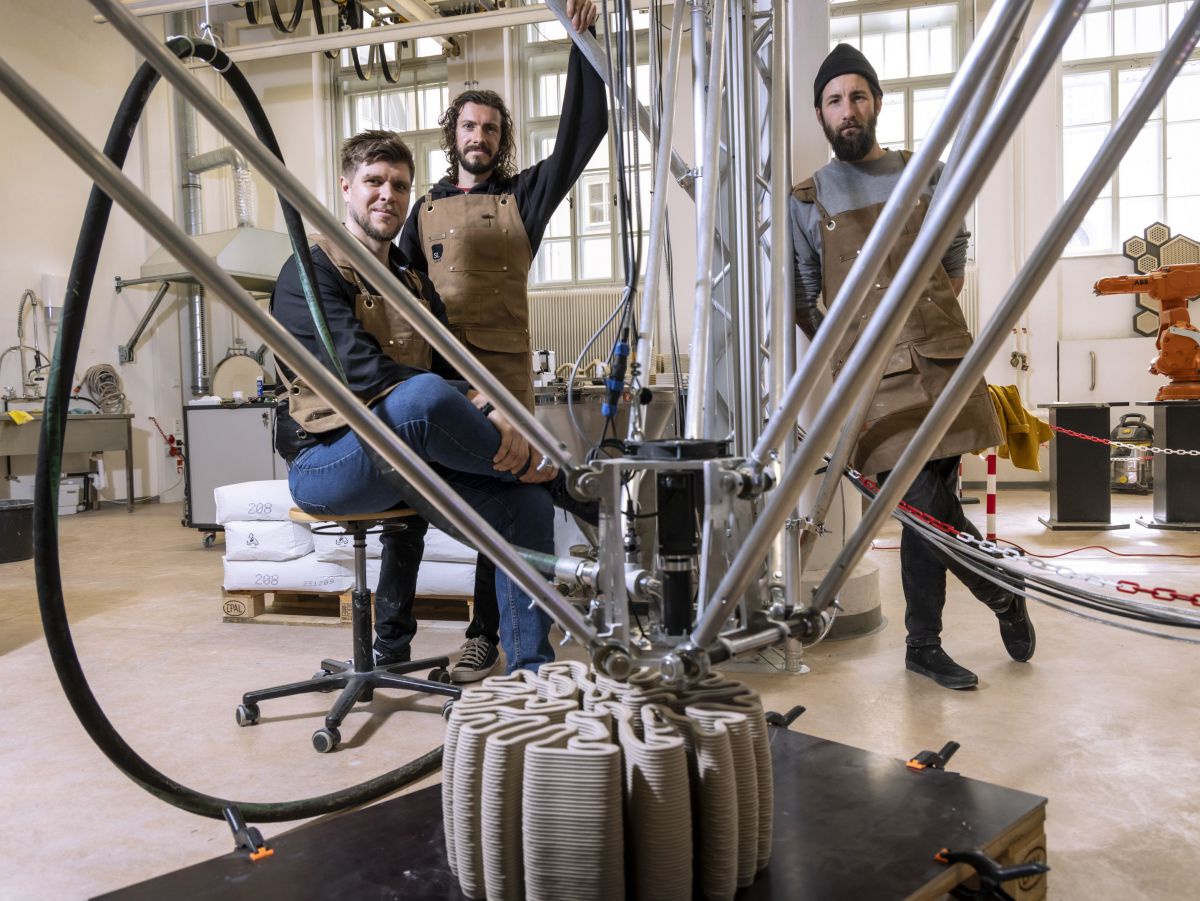 Aus den Düsen eines 3D-Druckers mit mehreren Armen wird Ton in einer korallenförmig geschlungenen Struktur auf ein schwarzes Podest gespritzt. Hinter dem Gerät stehen drei Männer mit braunen Arbeitsschürzen.