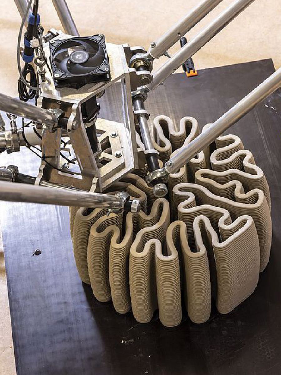 Aus den Düsen eines 3D-Druckers mit mehreren Armen wird Ton in einer korallenförmig geschlungenen Struktur auf ein schwarzes Podest gespritzt. 