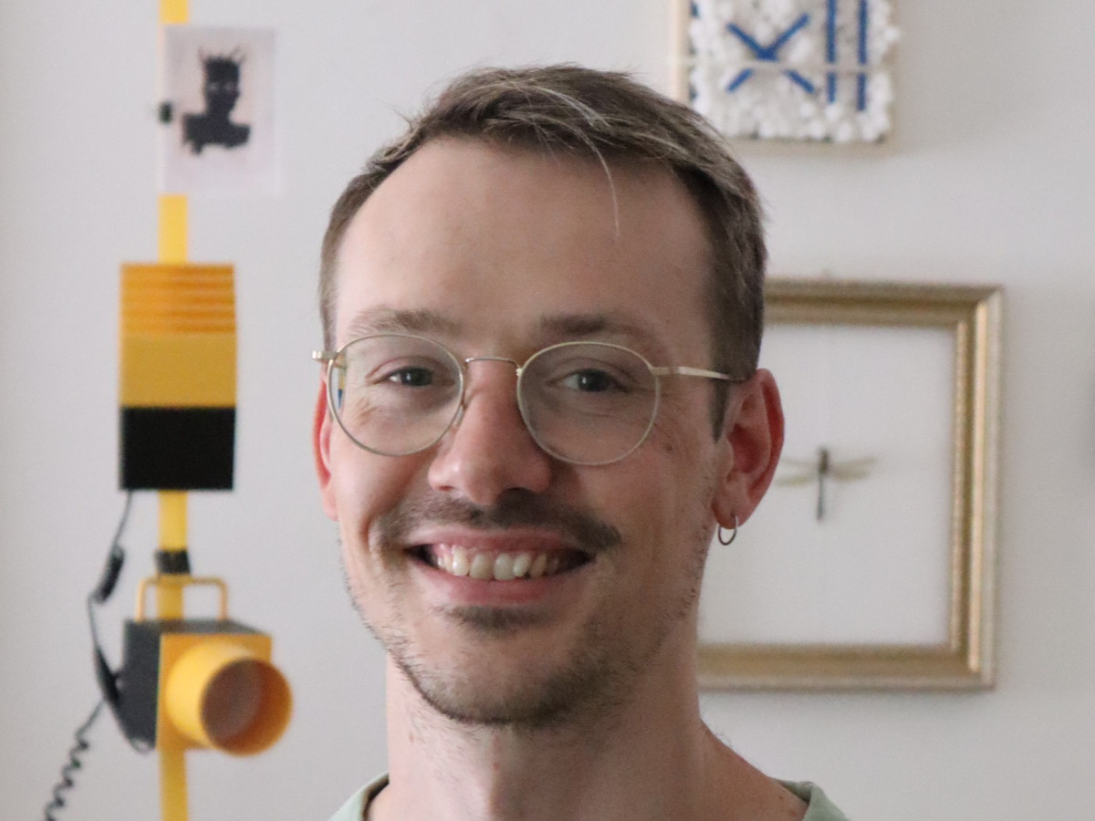 Dominik Scherrer: Ein junger Mann mit kurzem, hellem Haar, Dreitagesbart und Brille lächelt in die Kamera.