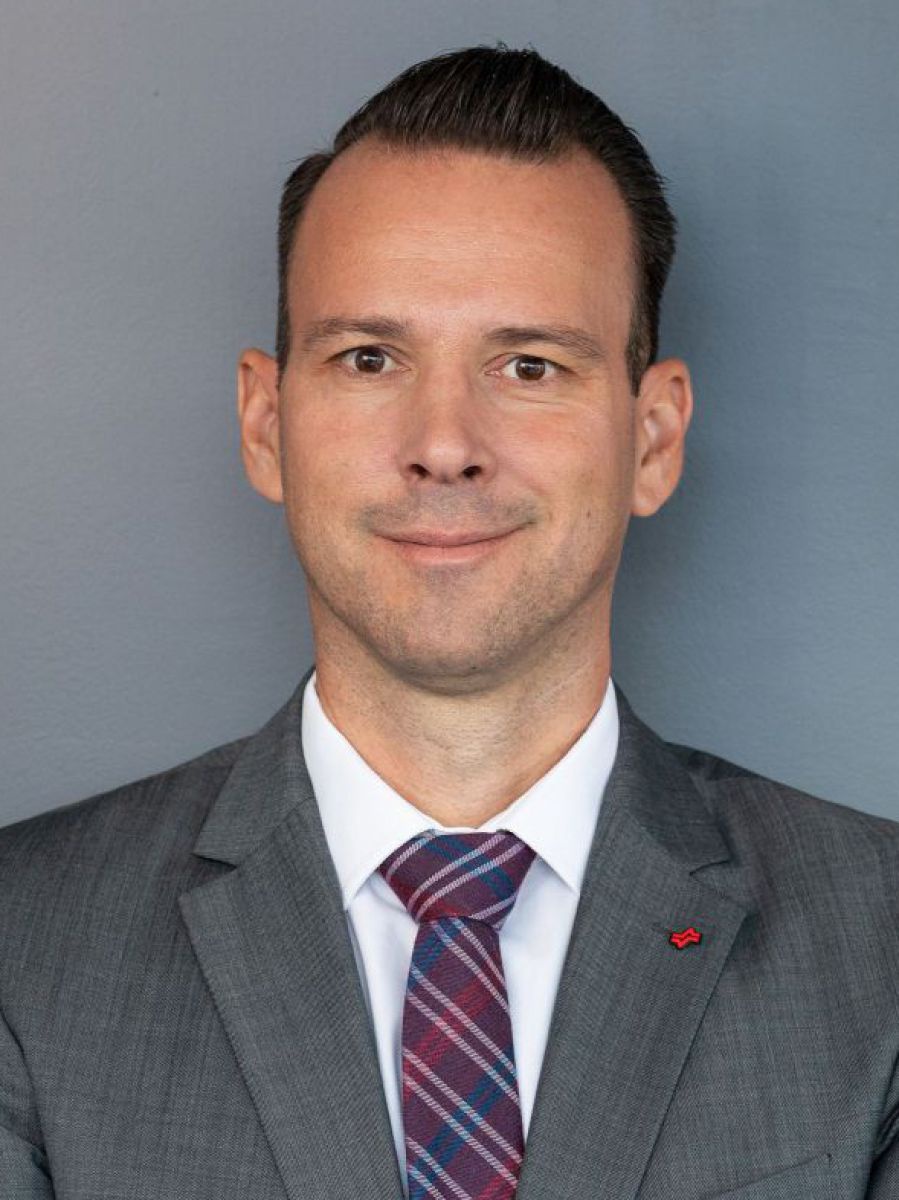 Portraitfoto eines Mannes um die Dreißig im grauen Sakko, weißem Hemd und Krawatte. Er steht, hat die Arme unter der Brust verschränkt und lächelt schmal.