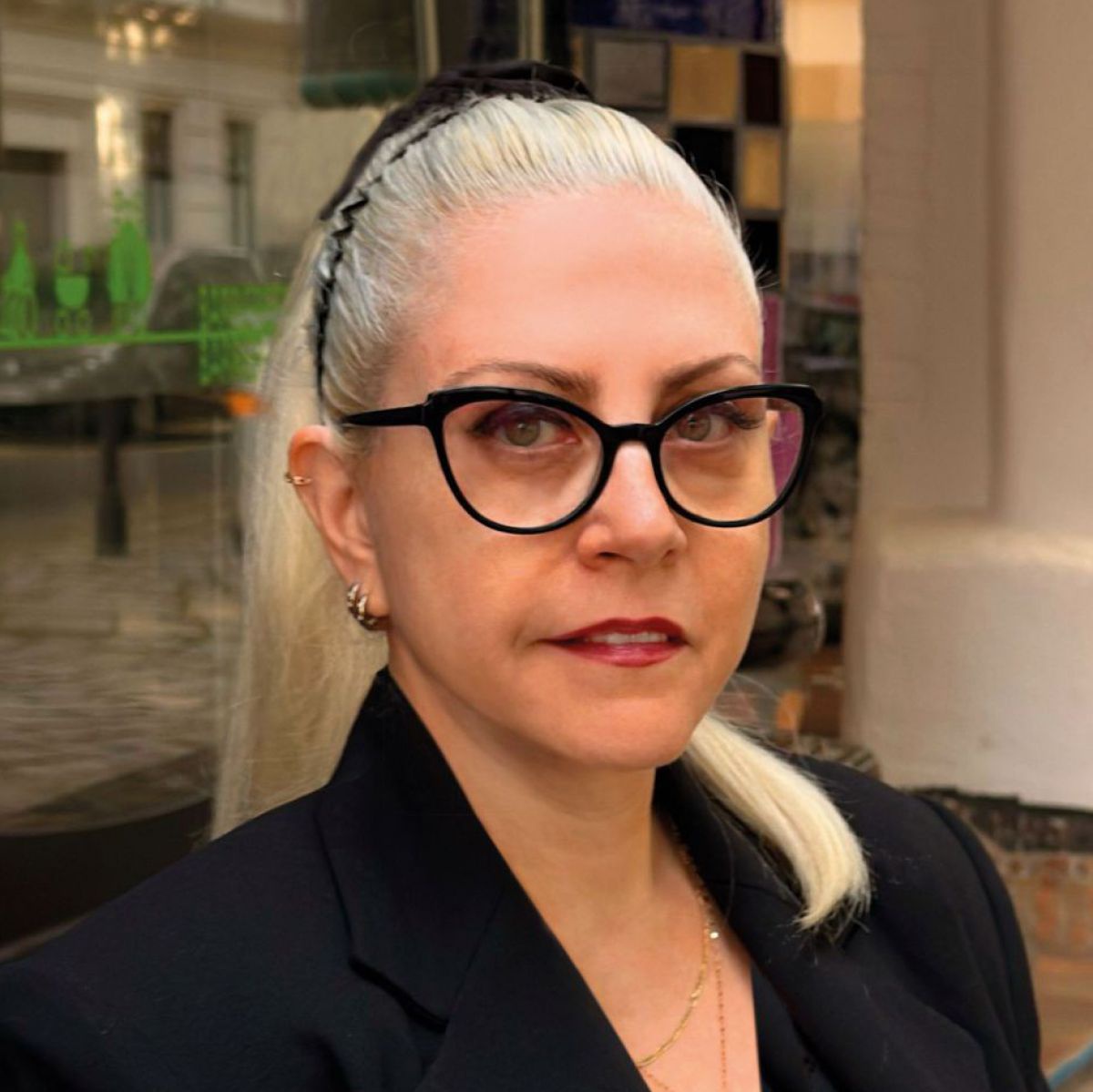 Eine Frau mit wasserstoffblondem Haar, das zu einem Pferdeschwanz gebunden ist, sie trägt eine Brille mit schwarzem, schmetterlingsflügelförmigen Rahmen, roten Lippenstift und einen schwarzen Blazer. Hinter ihr der Eingangsbereich des KunstHausWien mit ei