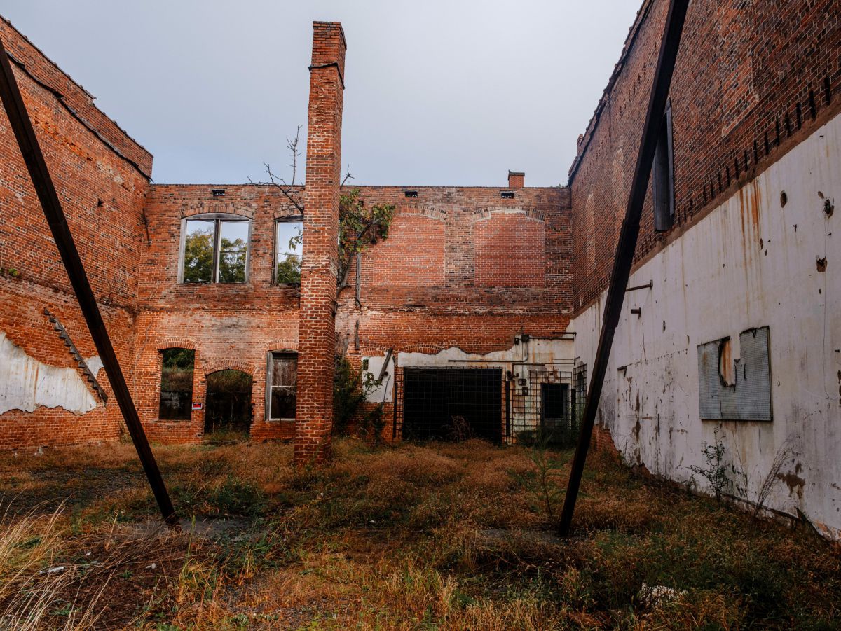 Blick in ein verfallenes Fabriksgebäude, ein gemauerter Schornstein steht noch in der Mitte, der einstige Boden ist mit Gras und Gestrüpp überwuchert.