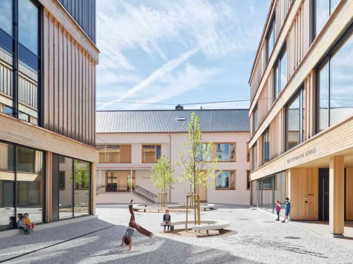 Blick in den Hof eines sanierten und durch neue Gebäude erweiterten Schulzentrums. Schüler*innen bewegen sich zwischen jungen Bäumen und Sitzgelegenheiten. Die neuen Gebäude sind aus hellem Holz und haben große Fensterflächen.