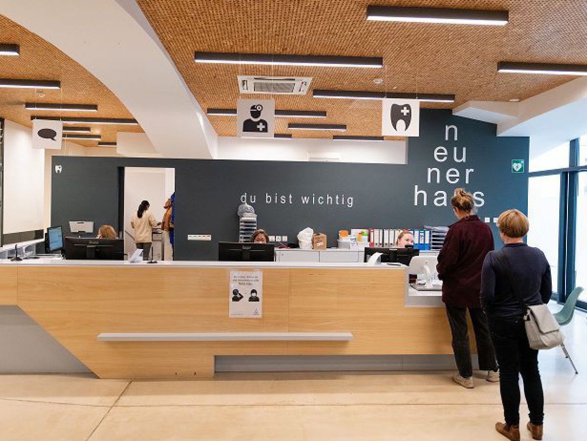 Blick ins Innere des neunerhaus Gesundheitszentrums: Zwei Menschen stehen angelstellt an der Anmeldungstheke. Der Raum ist lichtdurchflutet und mit viel hellem Holz ausgestattet.