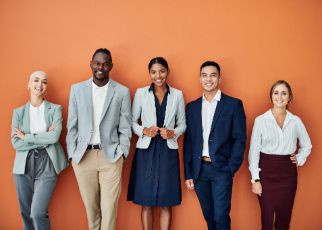 Ein Mitarbeiter*innen Team von 5 Personen verschiedenen Geschlechts, Ethnie und Körpergröße posiert vor einer orangen Wand.