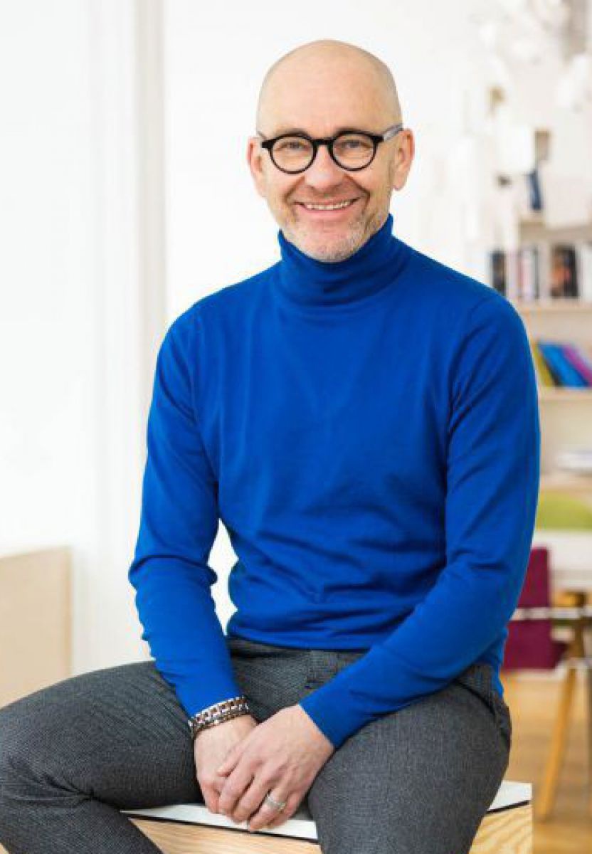 Ein Mann mit Glatze und grauem Bart sitzt auf einem würfelförmigem Holzmöbel in einem hellen Raum. Er trägt eine runde Brille mit dickem schwarzem Rahmen, einen blauen Rollkragenpullover und eine graue Slimfit Hose.