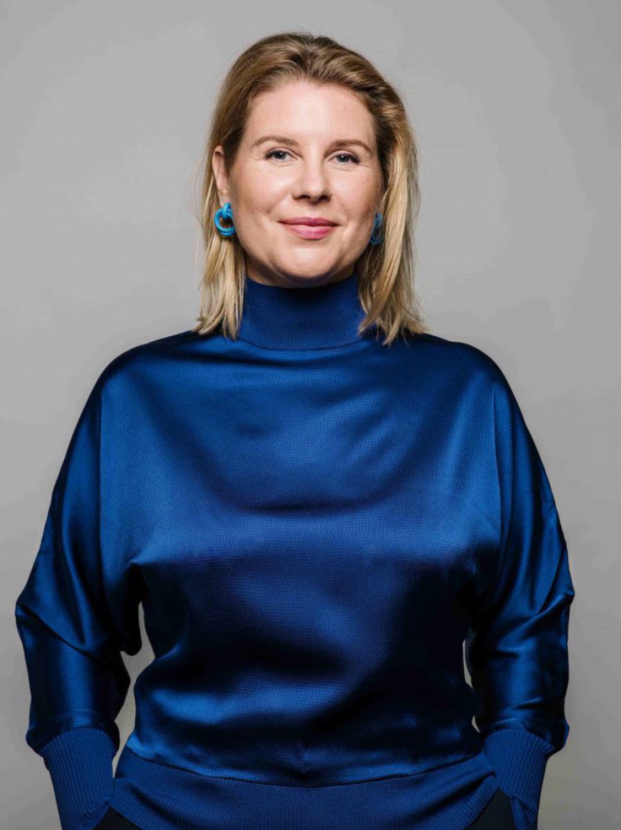 Eine Frau mit blondem, schulterlangem Haar posiert stehend vor grauem Hintergrund. Sie trägt eine dunkelblaue Seidenbluse mit langen Ärmeln und Turtelneck, ihre Hände hat sie in den Hosentaschen. Die hinter das rechte Ohr geklemmte Haarsträhne gibt den Bl