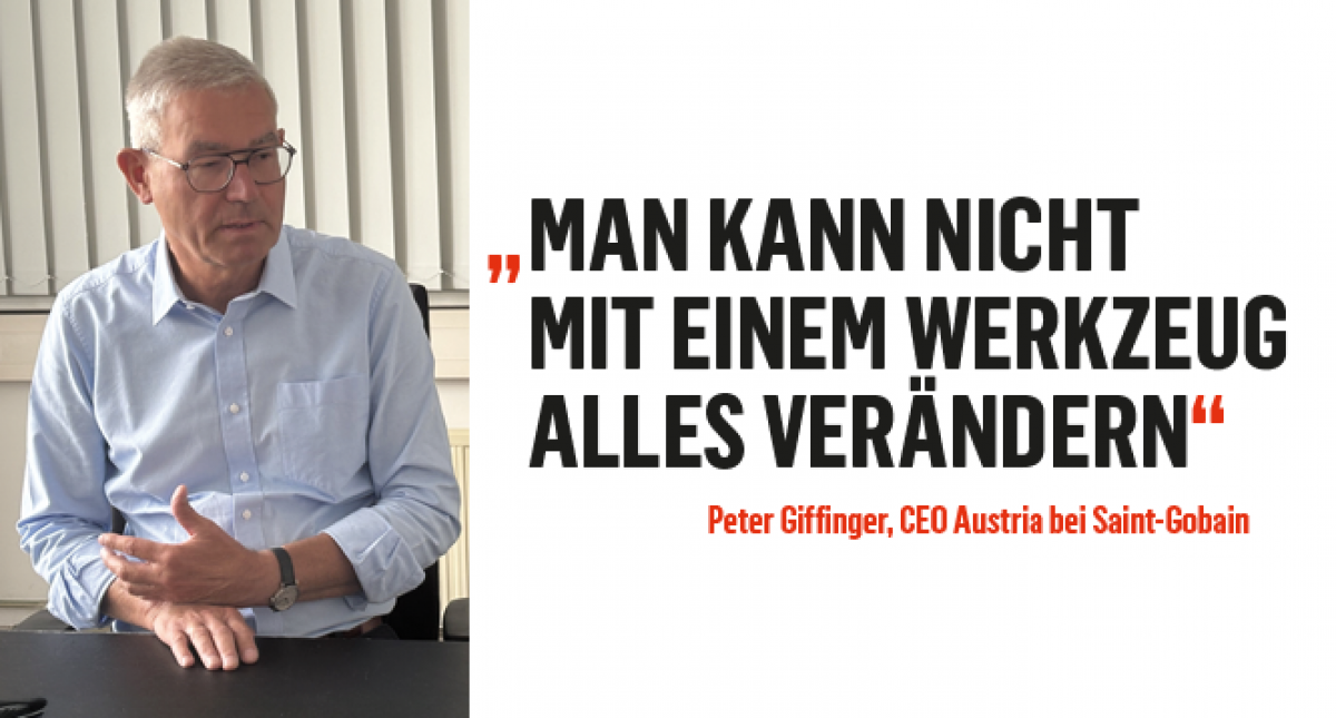 Mann um die 50 mit grauem Bürstenschnitt und Brille, trägt hellblaues Hemd mit offenem Kragen. Er sitzt an einem schwarzen Tisch und gestikuliert im Gespräch. Links vom Bild im Weißbereich steht: 