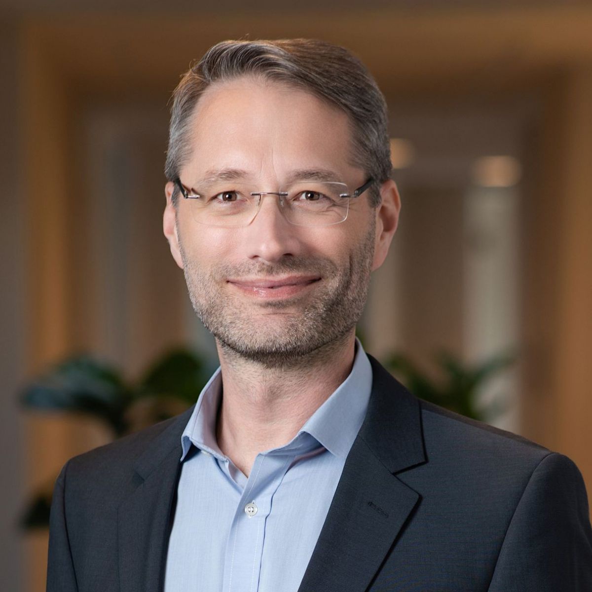 Portraitfoto mit verschwommenem Hintergrund: Ein Mann mit kurzem, braunem Haar, rahmenloser Brille und kurzem Bart lächelt mit geschlossenen Lippen. Sein langer Hals ragt aus dem offenen Kragen seines hellblauen Hemds, darüber trägt er ein dunkelblaues od