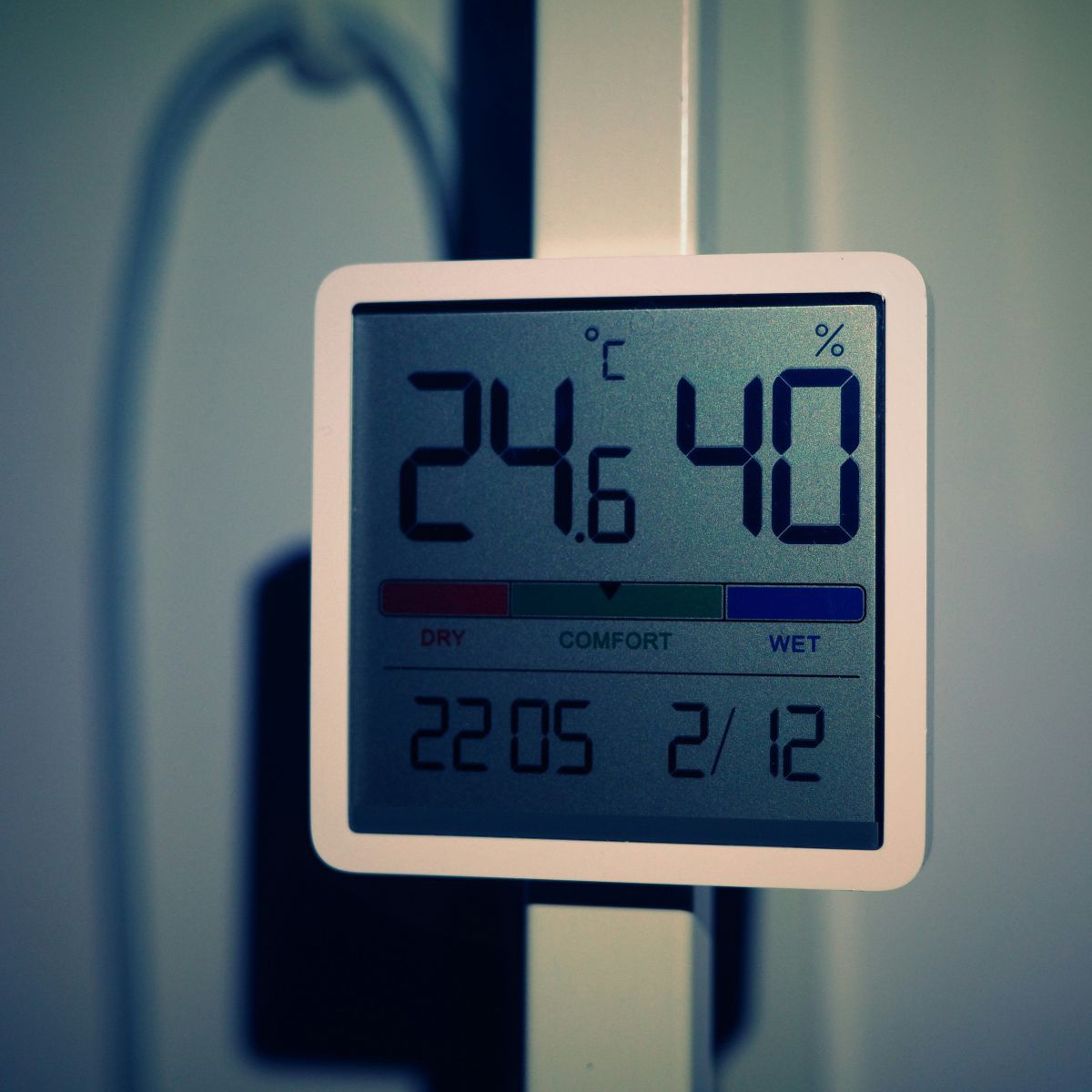 Digitale Anzeige, die Temperatur, Luftfeuchtigkeit, Datum und Uhrzeit anzeigt. Dahinter sieht man einen Kabelkanal und ein weißes Kabel an einer weißen Wand.