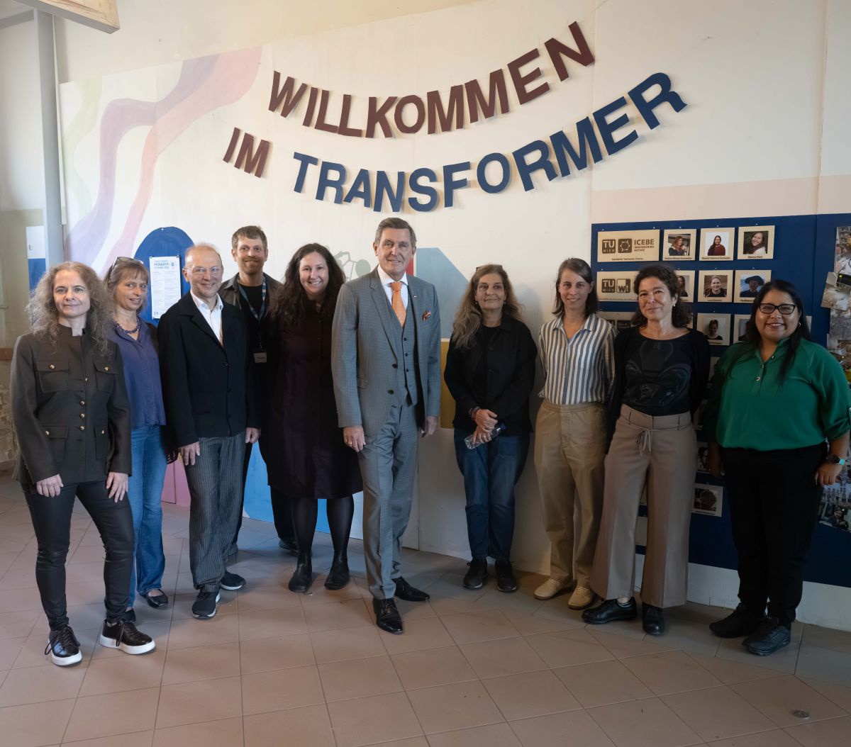Eine Gruppe von zehn Erwachsenen posiert für das Foto vor einer Wand, an der die Aufschrift 