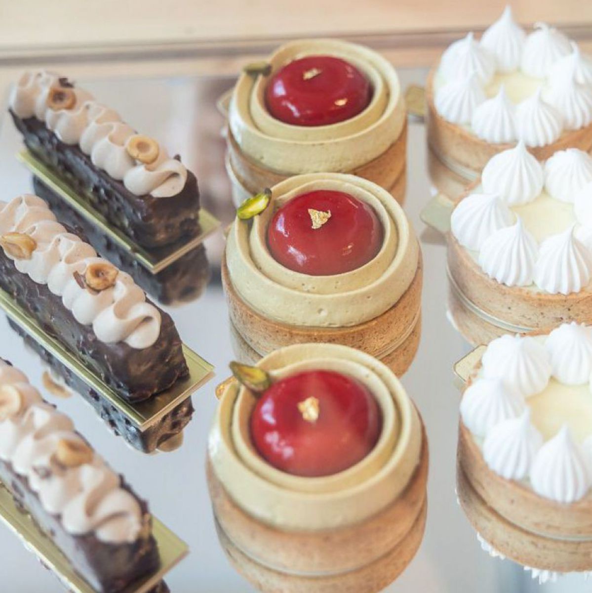 Drei verschiedene vegane Patisserie-Köstlichkeiten sind in Dreierreihen auf einem quadratischen Spiegel in einer Mehlspeisvitrine ausgestellt.
