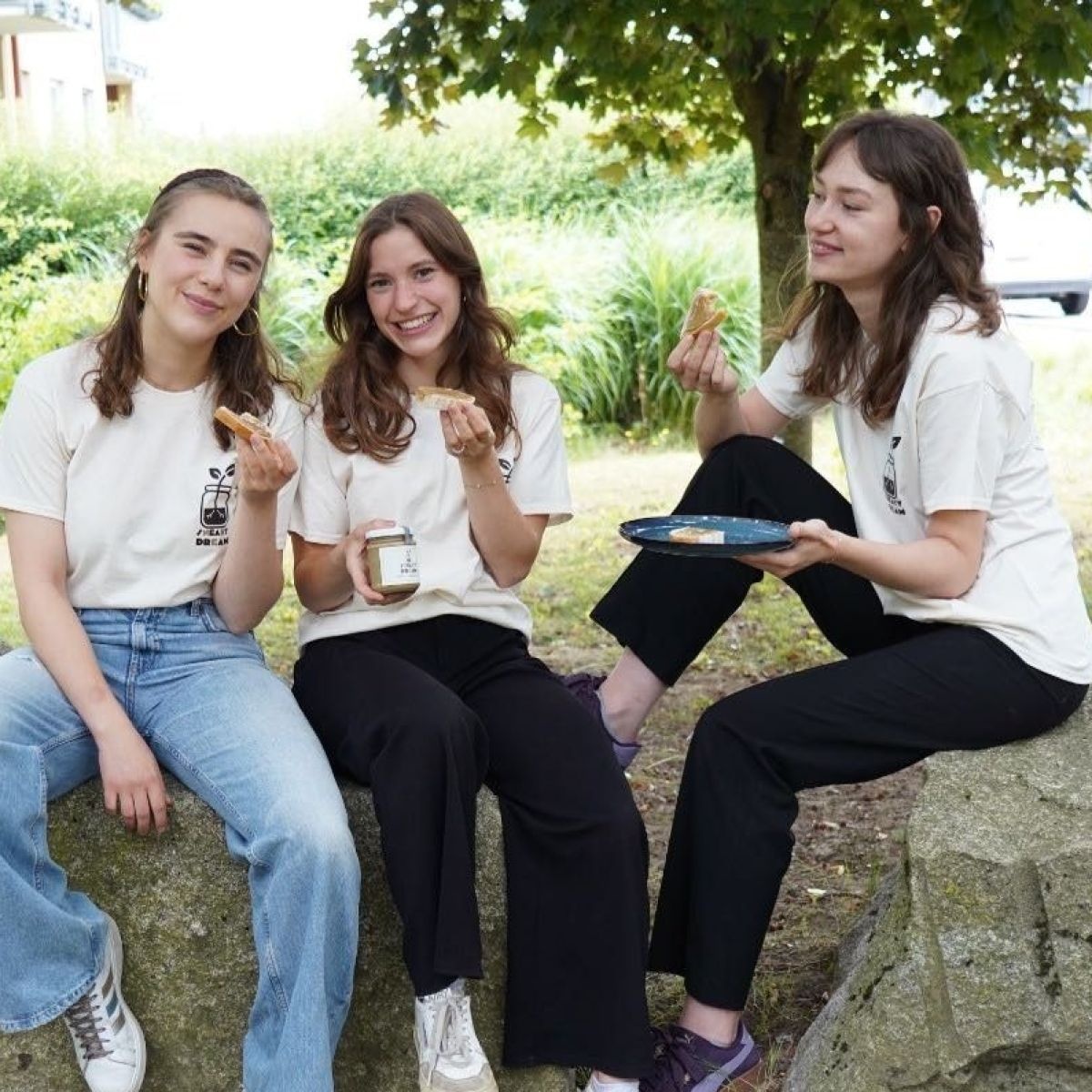 Drei junge Frauen mit langen Haaren sitzen in langen Hosen und T-Shirts auf großen Steinen und verkosten erdnussfreie Erdnussbutterbrote. Im Hintergrund ein Park und Gebäude.