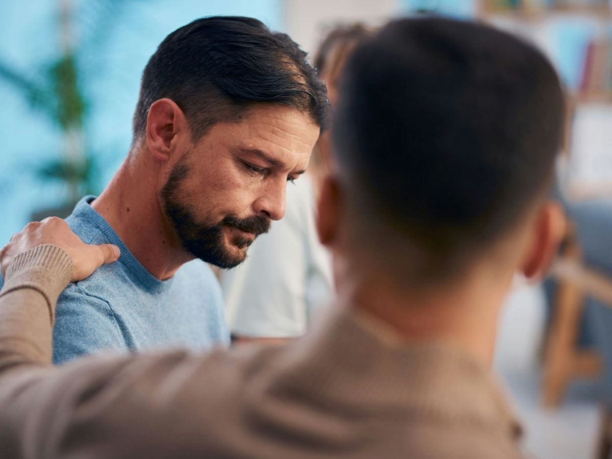 Ein Mann, den man nur verschwommen von hinten sieht, legt einem anderen Mann, der niedergeschlagen zu Boden blickt, mitfühlend die Hand auf die Schulter. Im Hintergrund sieht man verschwommen Tische, Stühle und eine Pflanze. 