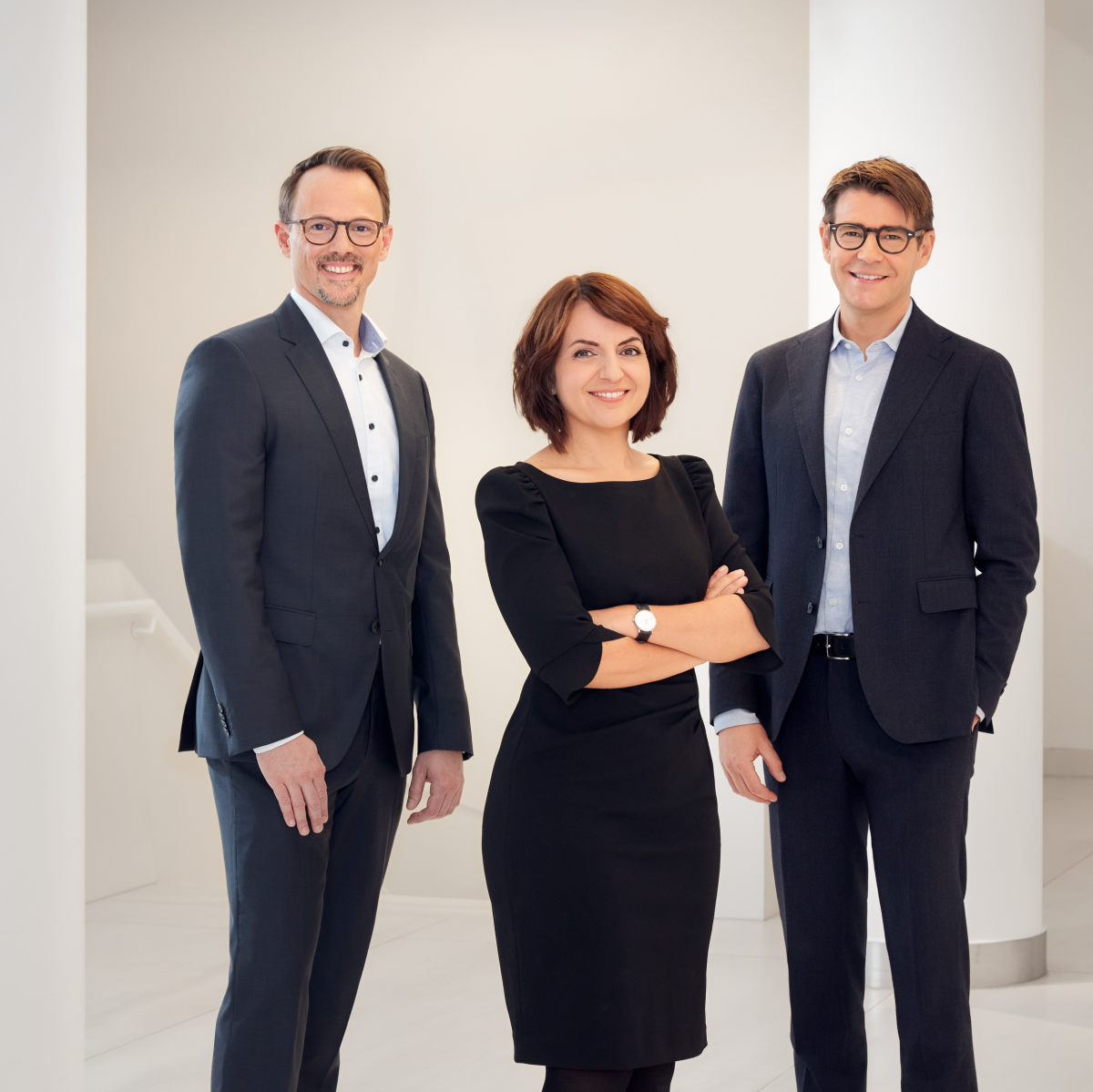 Gruppenfoto: Georg Jurceka, Anna Nowshad und Julian Mauhart posieren in dunkler Businesskleidung in einem weißen, kahlen Stiegenhaus. 