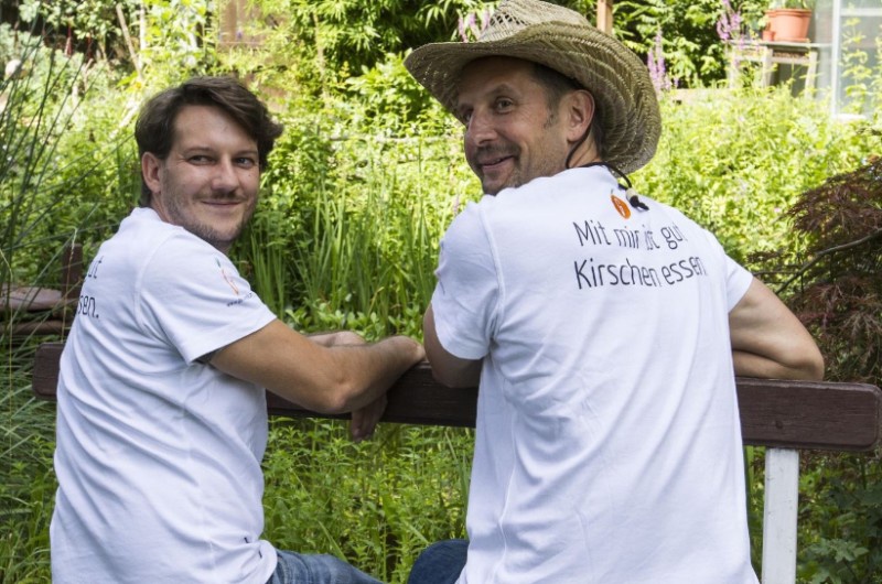 Zwei Männer mit weißen T-Shirts vor einem grünen Garten