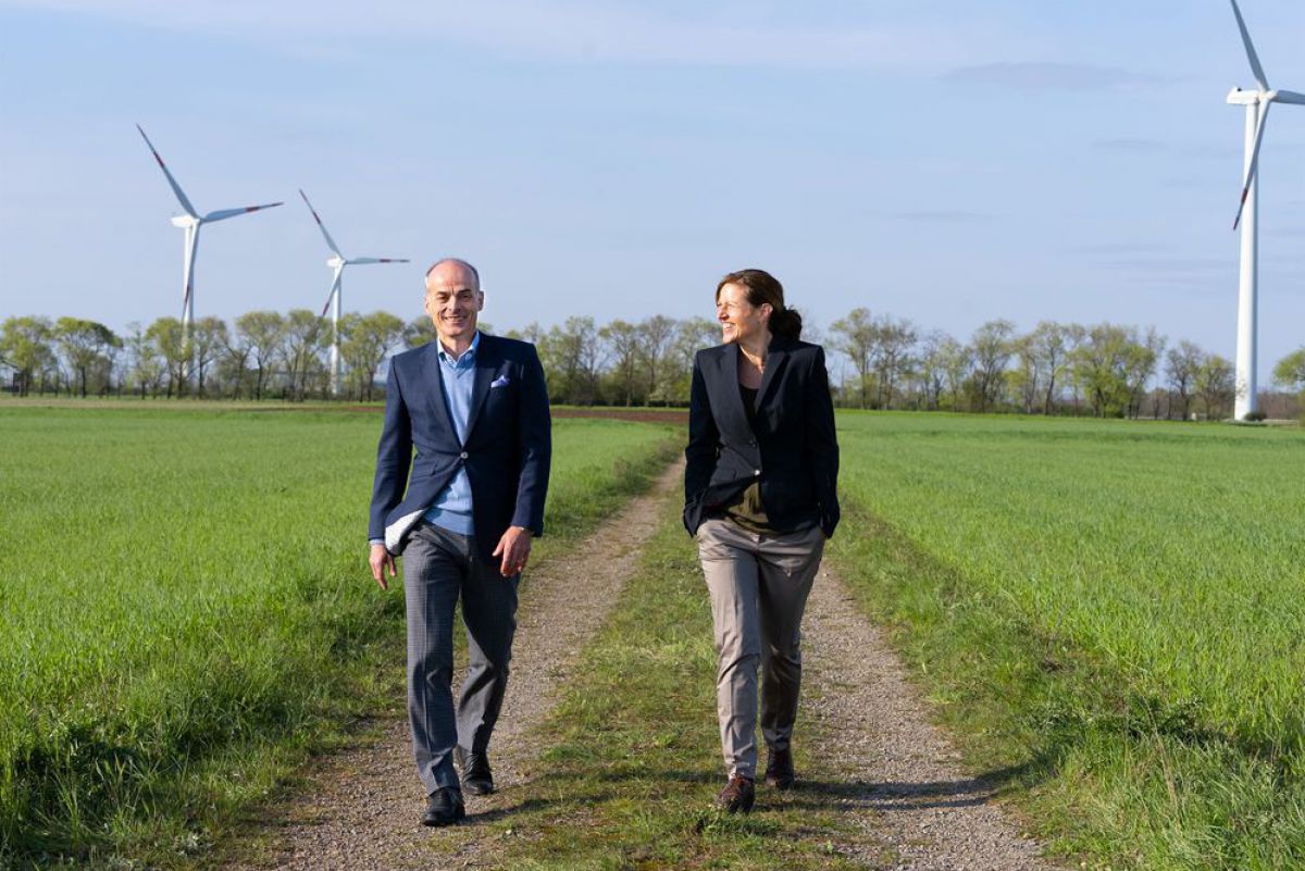 Streibl und Aichberger gehen einen Feldweg entlang. Im Hintergrund sieht man drei Windräder.