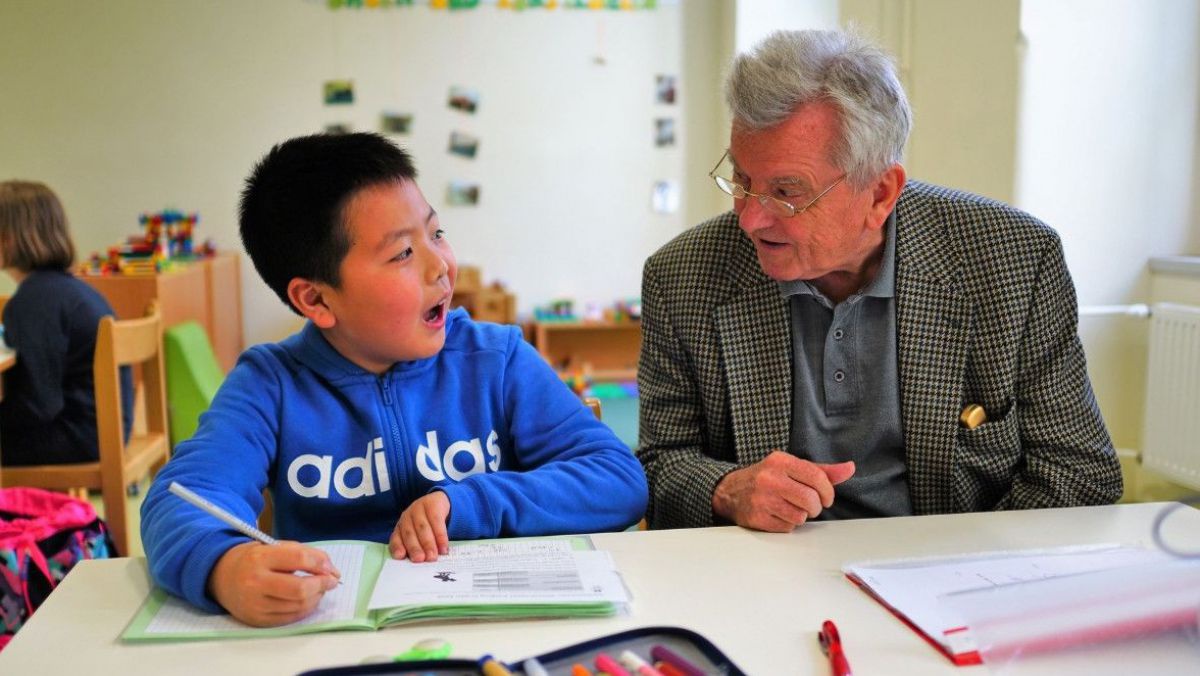 Ein Volkssschüler sitzt mit Heft und Bleistift an einer Schulbank, neben ihm ein älterer Herr, der ihn unterstützt, die beiden scheinen sich angeregt zu unterhalten.