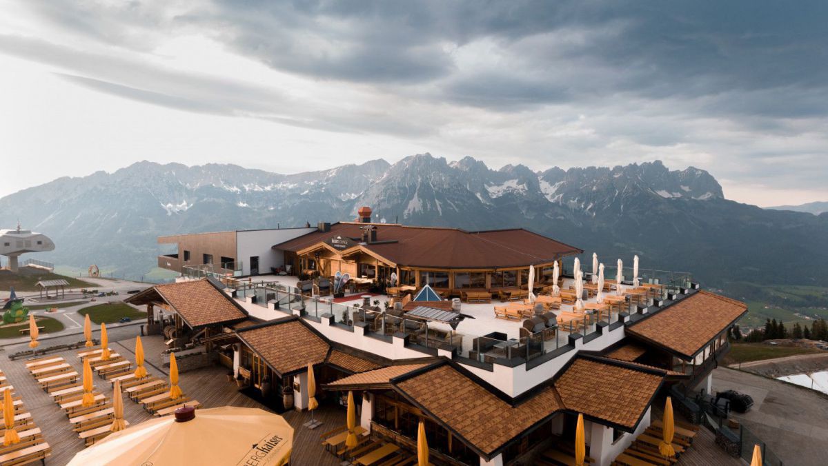 Ein Restaurant, hoch in den Bergen gelegen. Im Hintergrund eine Gebirgskette unter dunkel bewölktem Himmel.