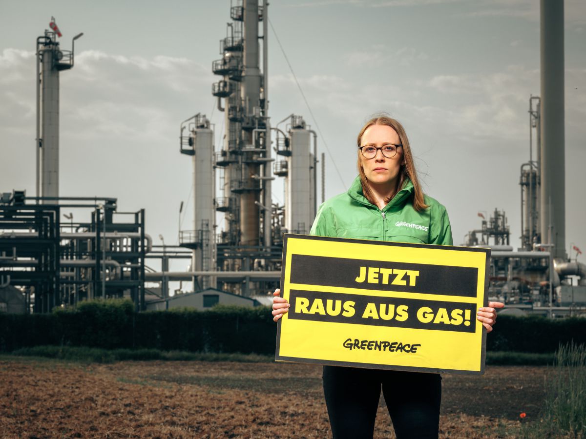 Eine Frau in grüner Greenpeace-Jacke steht vor einem Gaswerk und hält ein gelbes Schild mit der Aufschrift: Jetzt raus aus Gas!