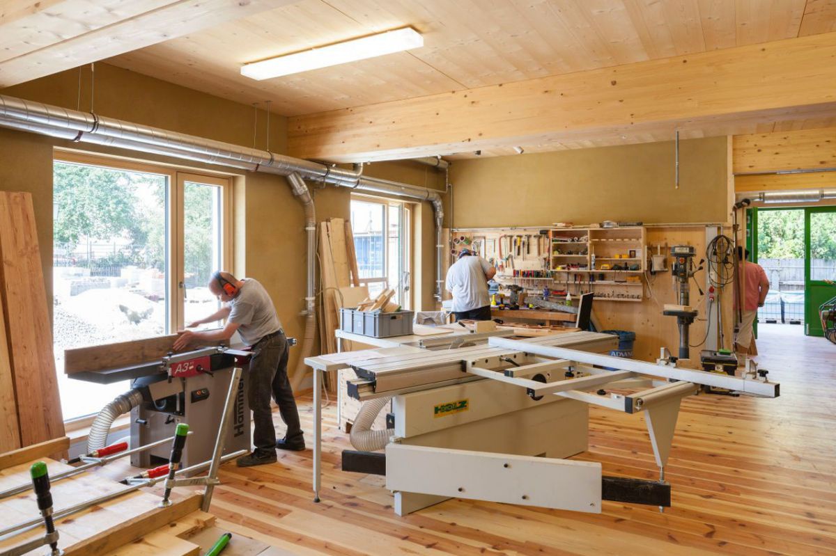 Auf dem Foto sieht man eine Tischlereiwerkstätte mit den entspechenden Maschinen in der zwei Männer arbeiten.