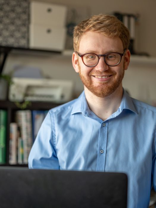 Ein junger Mann mit Bart und Brille an seinem Laptop am Schreibtisch lächelt in die Kamera, im Hintergrund Büroregale.