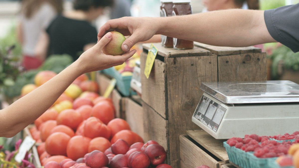 Über einem Marktstand mit Früchten sieht man den Arm des Verkäufers, der der Kundin eine Frucht in die Hand legt.
