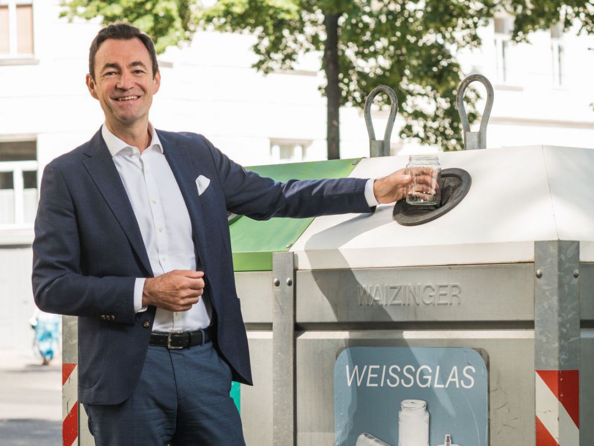 Dr. Harald Hauke, Geschäftsführer der Austria Glas Recycling GmbH steht lächelnd im graublauen Sakko bei einem Altglassammelcontainer und hält mit der linken Hand ein leeres Glasgebinde über die Einwurföffnung des Weissglascontainers.