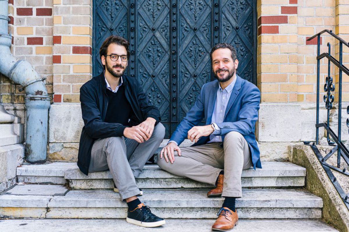 Thomas Ebenstein und Armand Colard sitzen auf den Steinstufen vor einem Backsteingebäude mit altem schmiedeeisenem Eingangstor.